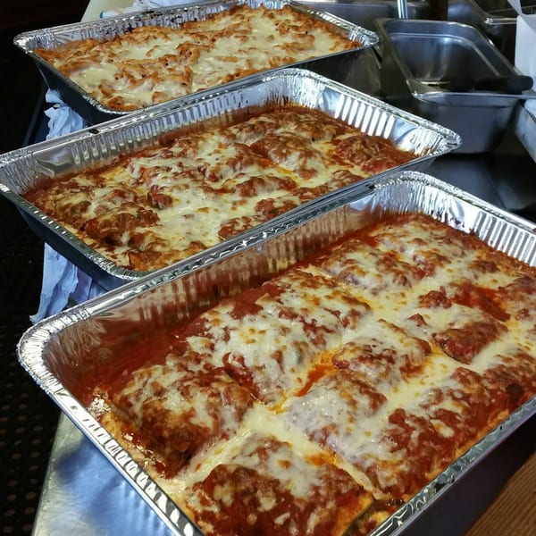Three aluminum trays filled with baked ziti and lasagna, topped with melted cheese and sauce.