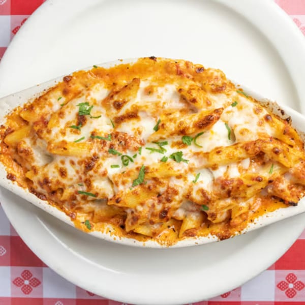 Baked penne pasta with melted cheese and parsley in a white dish on a red and white checkered tablecloth.