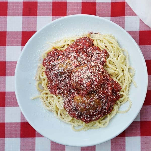 Spaghetti and meatballs with red sauce and parmesan cheese on a white plate over a red and white checkered tablecloth.