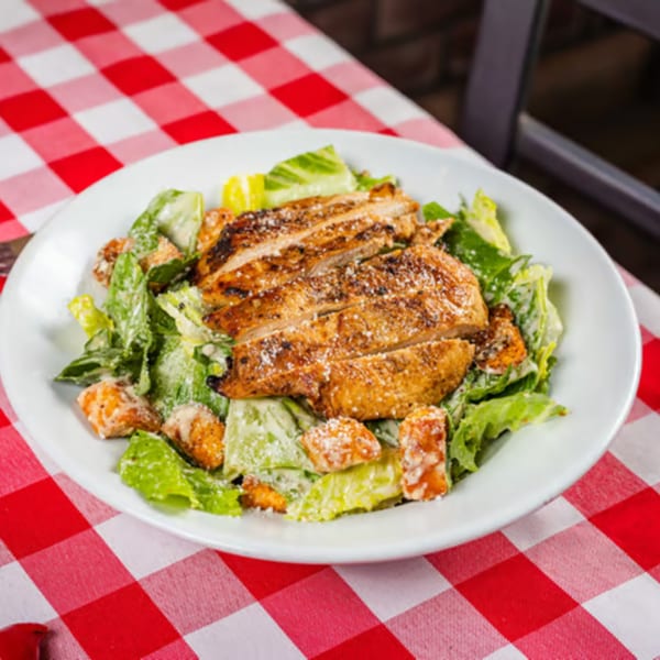 Grilled chicken Caesar salad with croutons and dressing on a white plate, on a red and white checkered tablecloth.