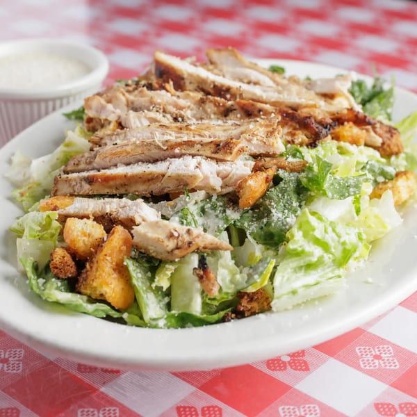 Chicken Caesar salad with croutons and Parmesan cheese on a white plate, on a red and white checkered tablecloth.