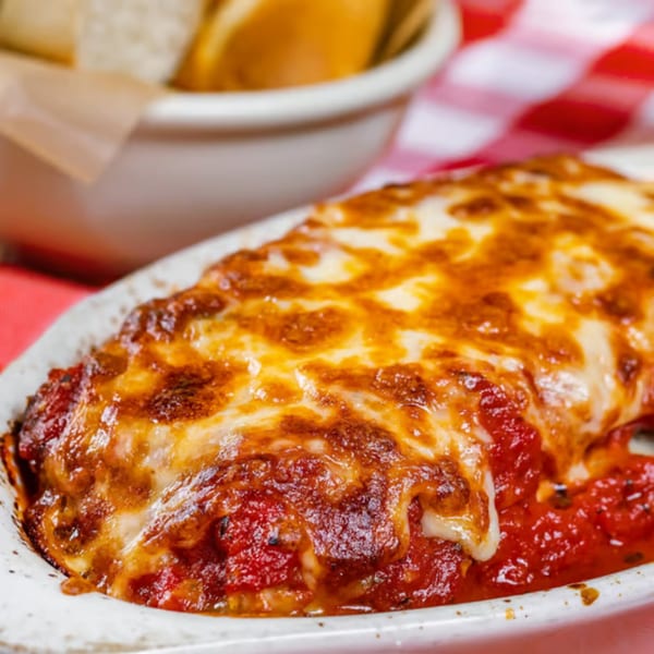 Baked pasta dish with melted, bubbly cheese and rich red sauce, with bread in the background.
