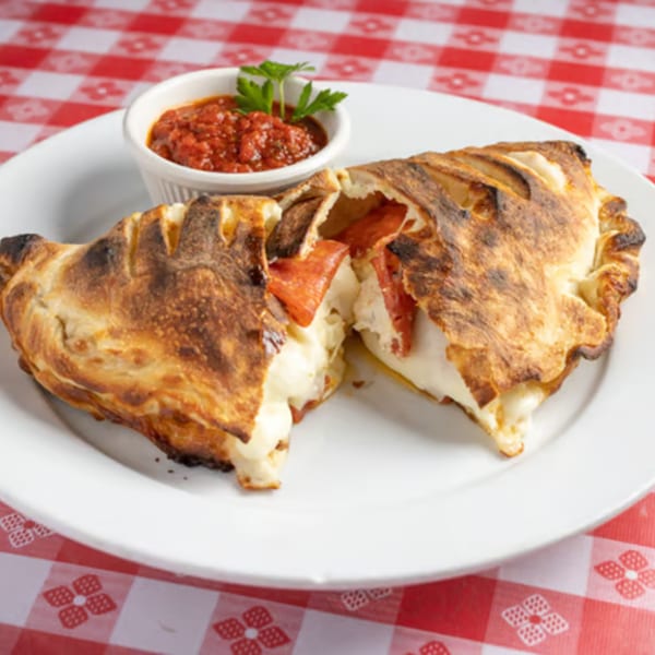 A golden-brown pepperoni and cheese calzone, split open, with a side of marinara sauce on a red and white checkered tablecloth.
