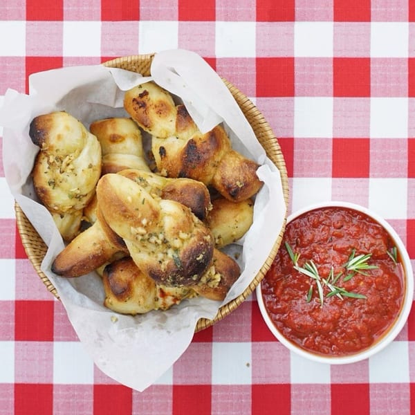 Garlic knots in a basket with a side of marinara sauce on a red and white checkered tablecloth.