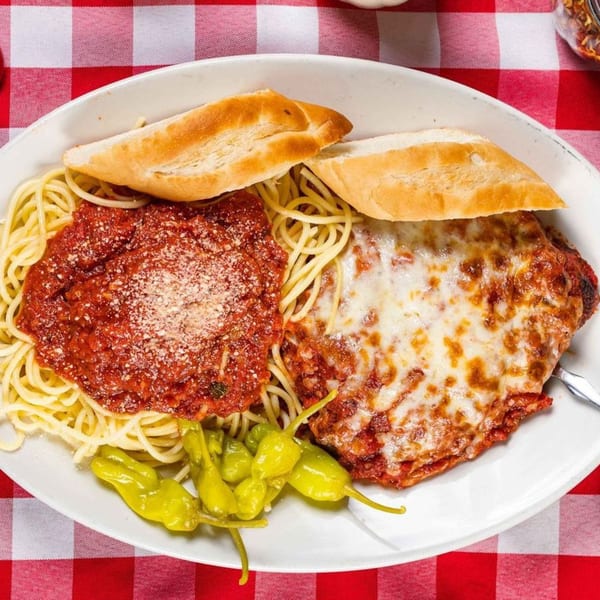 Spaghetti with red sauce, chicken parmesan, bread, and peppers on a white plate over a red and white checkered tablecloth.