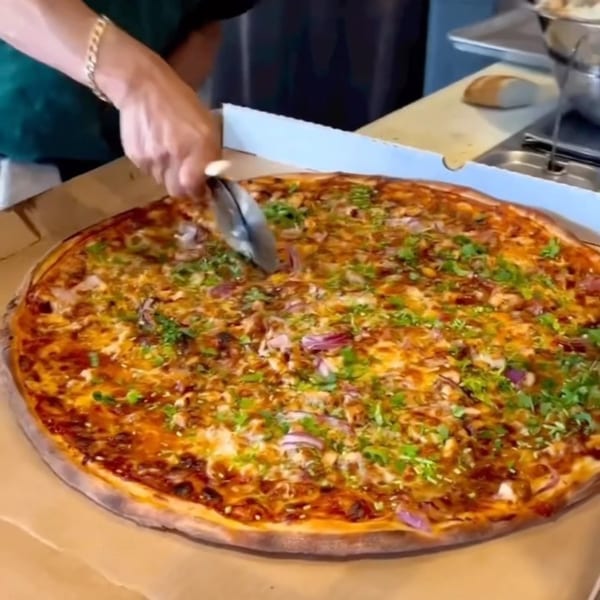 A person slices a large, freshly baked Enzo's Pizzeria pizza topped with herbs and onions.