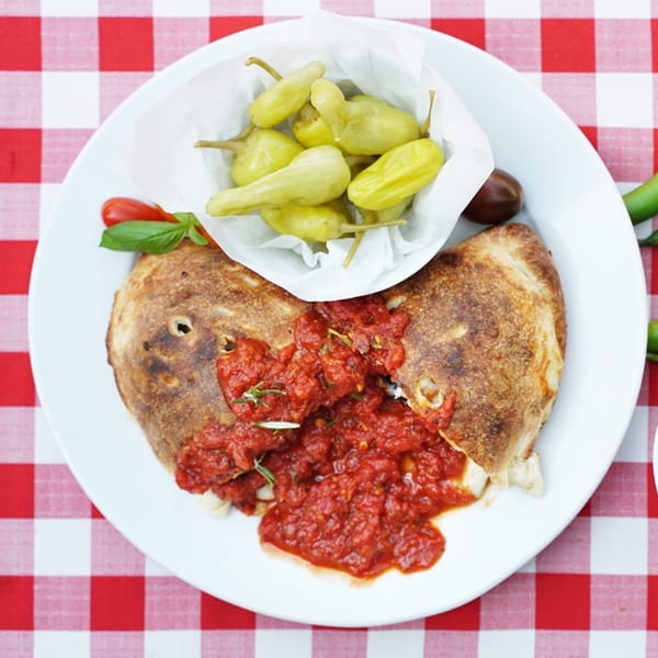 Enzo's Pizzeria calzone with marinara sauce and a side of pickled peppers on a red and white checkered tablecloth.
