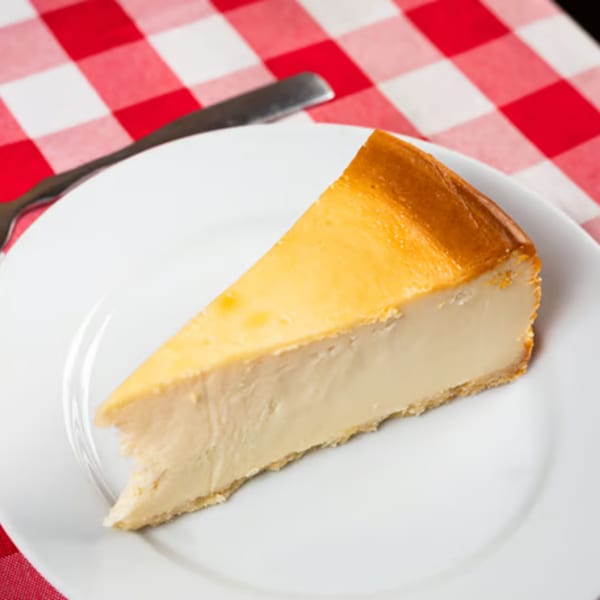 Slice of classic cheesecake on a white plate with a red and white checkered tablecloth.